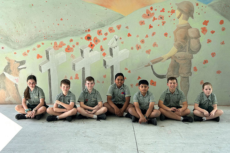 School students seated in front of a remembrance mural featuring red poppies, military graves and a First World War soldier.